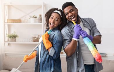 A young woman and man leaning on each other smiling and holding a duster and a brush