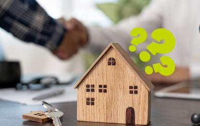 A wooden model of a house on a table next to a keychain with a silver key and a model house keyring on it. There are question marks around the model house and a handshake in the background