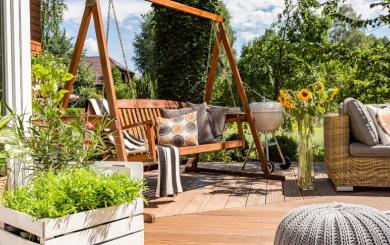 A wooden decking area with a swing seat, sunflowers, a barbecue and more plants 