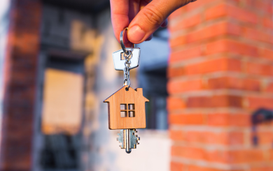A house key with a home shaped wooden key ring attached are being held by two fingers. In the blurred background you can see a red bricked home.