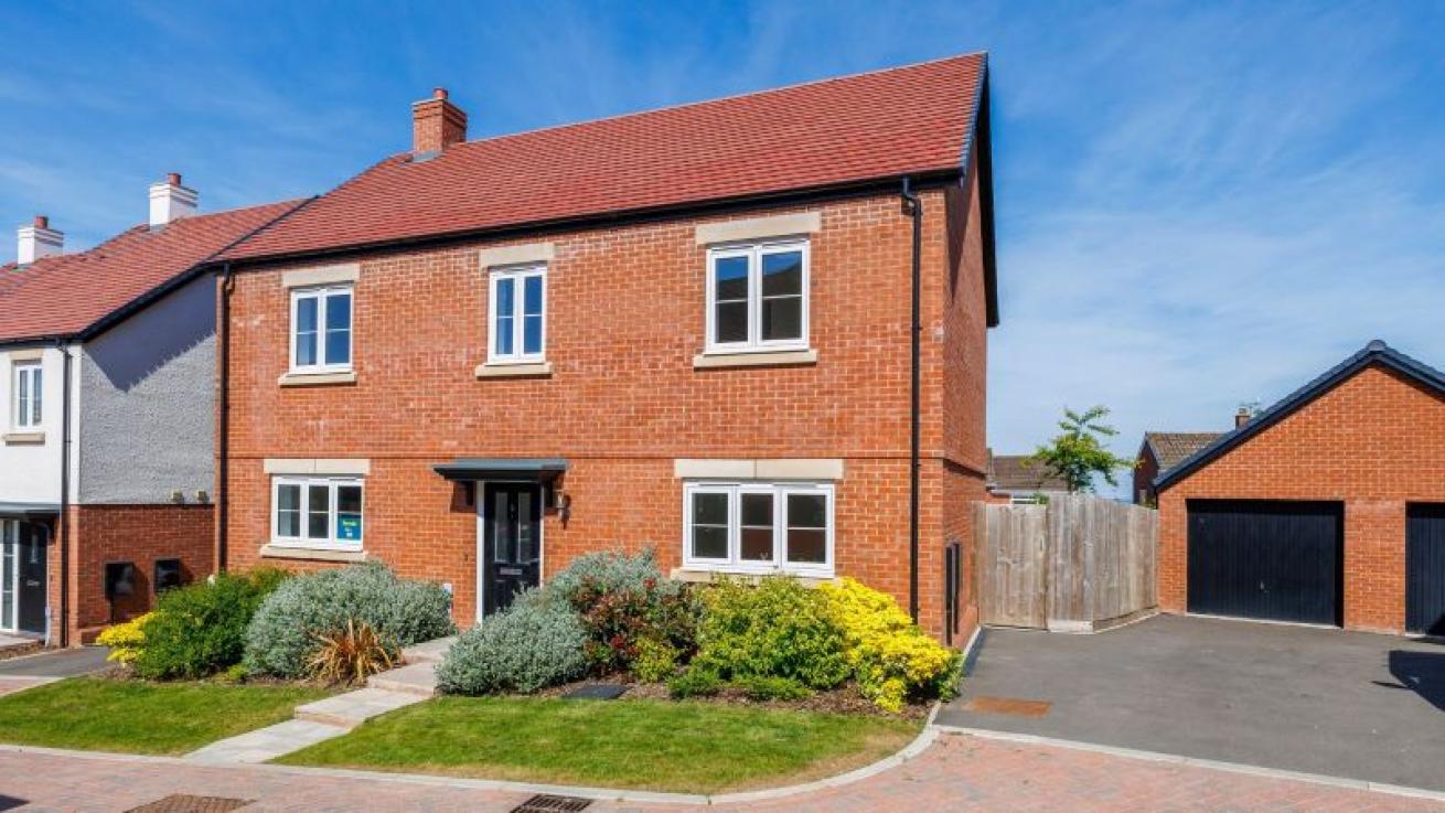 Modern two-story detached brick home with red-tiled roof, white-framed windows, landscaped front garden, paved path, and driveway leading to a detached garage.