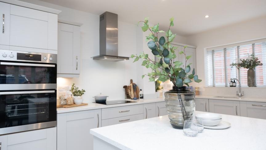 Interior of a clean and tidy white kitchen with cream cabinets. In the foreground is a kitchen island on which is a vase with foliage in and some crockery next to it