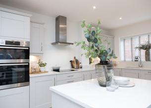 Interior of a clean and tidy white kitchen with cream cabinets. In the foreground is a kitchen island on which is a vase with foliage in and some crockery next to it